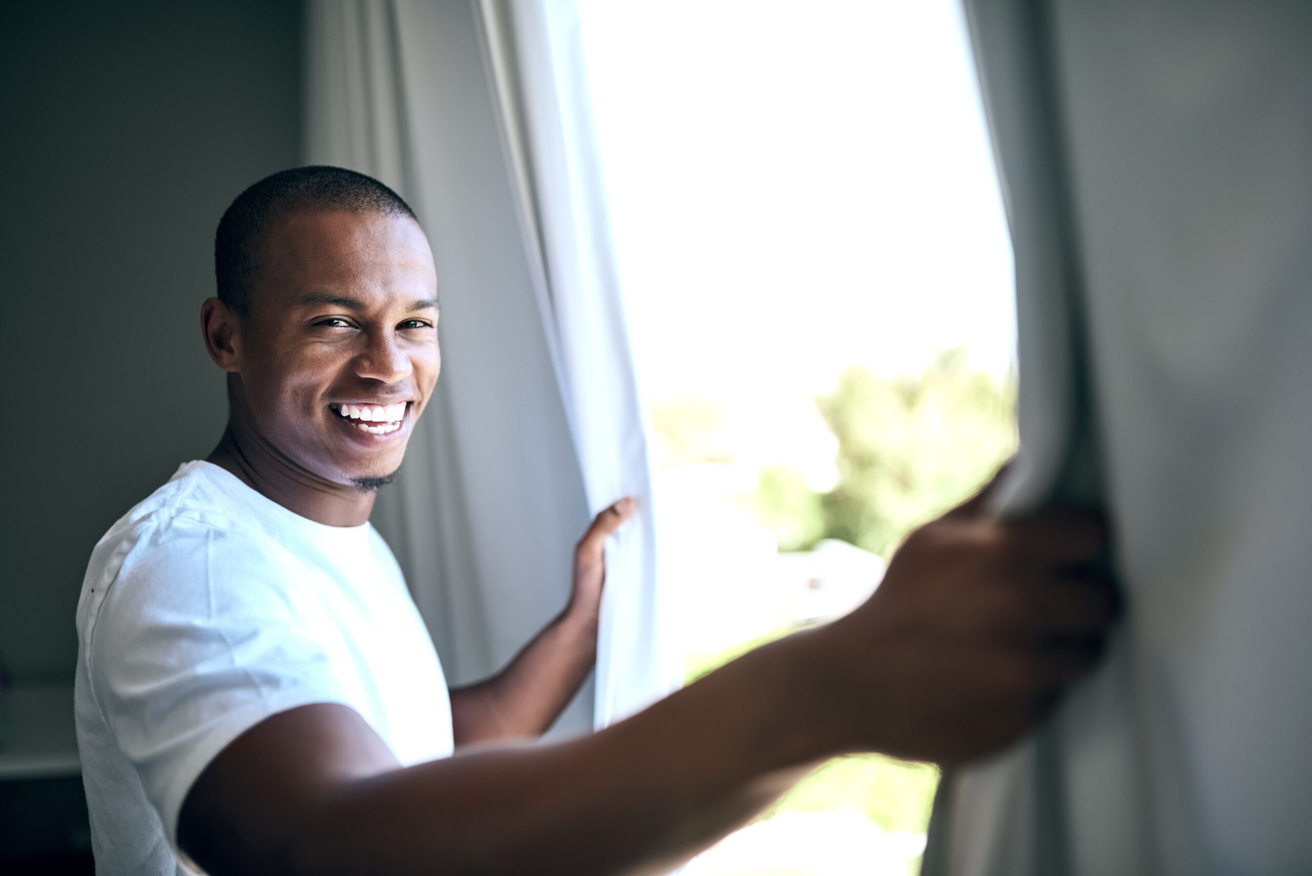 Male waking up in a sober living home smiling while holding curtains
